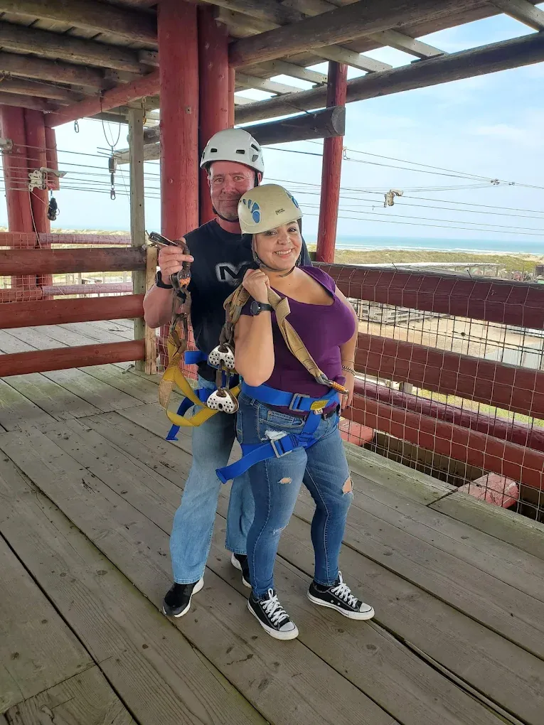 Two people in ziplining harnesses and helmets on the tower platform at South Padre Island Adventure Park